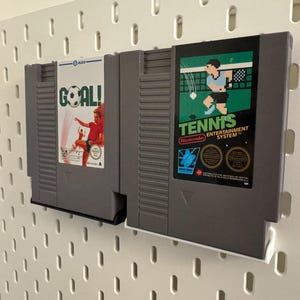 Pode incluir: Dois cartuchos de jogo cinzentos do Nintendo Entertainment System. Um apresenta um jogador de futebol com a palavra "GOAL!" e o outro mostra um jogador de t&eacute;nis com a palavra "TENNIS". Os cartuchos est&atilde;o montados numa placa perfurada branca.