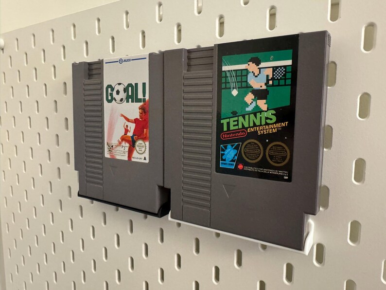 Pode incluir: Dois cartuchos de jogo cinzentos do Nintendo Entertainment System. Um apresenta um jogador de futebol com a palavra "GOAL!" e o outro mostra um jogador de t&eacute;nis com a palavra "TENNIS". Os cartuchos est&atilde;o montados numa placa perfurada branca.