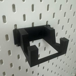 K&ouml;nnte beinhalten: Schwarzer Wandhalter aus Kunststoff f&uuml;r eine Lochwand. Der Halter hat eine rechteckige Form mit einer kleinen Lippe an der Vorderseite.