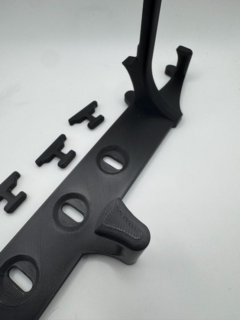 May include: A black 3D-printed stand with a long, flat base featuring three oval cutouts. The stand has a curved support and several small, H-shaped pieces. The items are arranged on a white surface.