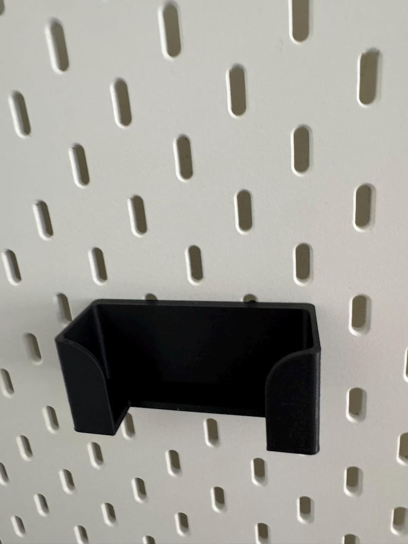 Pu&ograve; includere: Porta telefono in plastica nera montato su una parete in pannello forato bianco.