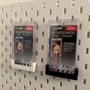 以下が含まれることがあります： 白と黒のUltra Pro Booster Pack One-Touchケースが2つ、白いペグボードに取り付けられています。各ケースには、アニメスタイルのイラストが描かれたカードが入っています。ケースはUV保護、ダイヤモンドコーナー、マグネットクロージャーを備えています。