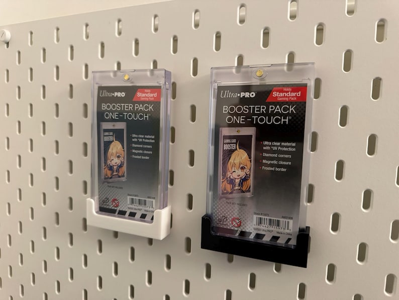 以下が含まれることがあります： 白と黒のUltra Pro Booster Pack One-Touchケースが2つ、白いペグボードに取り付けられています。各ケースには、アニメスタイルのイラストが描かれたカードが入っています。ケースはUV保護、ダイヤモンドコーナー、マグネットクロージャーを備えています。