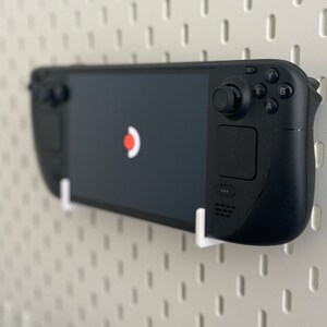 Puede incluir: Una consola de juegos port&aacute;til negra con una pantalla, botones y una palanca de mando. La consola est&aacute; montada en un tablero de clavijas blanco.