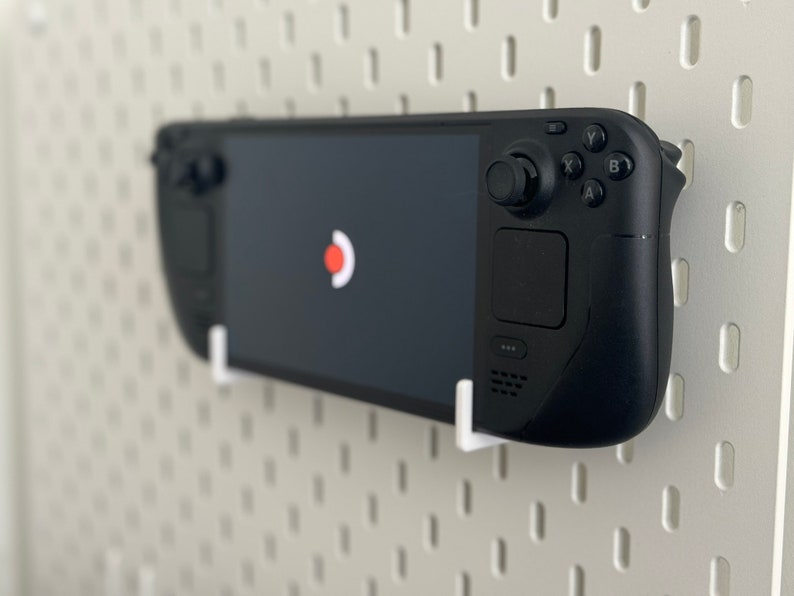 Puede incluir: Una consola de juegos port&aacute;til negra con una pantalla, botones y una palanca de mando. La consola est&aacute; montada en un tablero de clavijas blanco.
