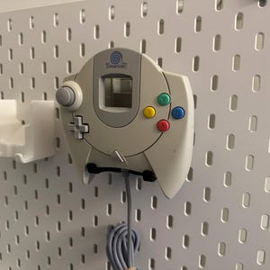 K&ouml;nnte beinhalten: Ein cremefarbener Dreamcast-Controller mit farbigen Kn&ouml;pfen, montiert auf einer wei&szlig;en Lochwand. Der Controller hat einen grauen Analog-Stick, ein Steuerkreuz und ein Logo. Links befindet sich ein wei&szlig;er Halter.