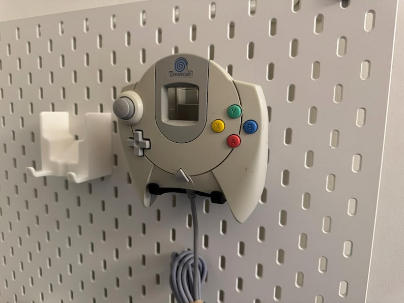 K&ouml;nnte beinhalten: Ein cremefarbener Dreamcast-Controller mit farbigen Kn&ouml;pfen, montiert auf einer wei&szlig;en Lochwand. Der Controller hat einen grauen Analog-Stick, ein Steuerkreuz und ein Logo. Links befindet sich ein wei&szlig;er Halter.