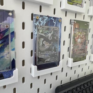 K&ouml;nnte beinhalten: Eine wei&szlig;e Lochwand mit mehreren durchsichtigen Plastikkartenhaltern, die verschiedene Pok&eacute;mon-Sammelkarten zeigen. Die Karten sind in verschiedenen Farben und zeigen verschiedene Pok&eacute;mon-Charaktere. Die Karten sind in einem Raster auf der Lochwand angeordnet.
