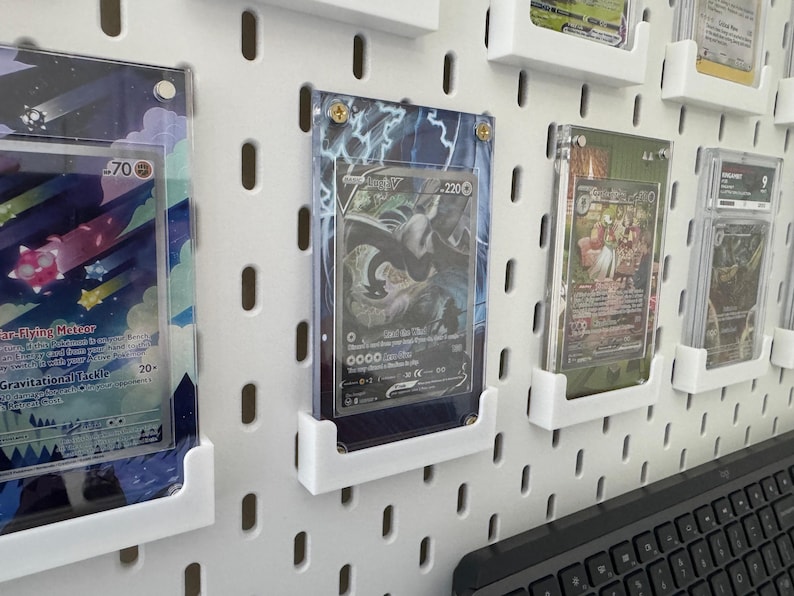 K&ouml;nnte beinhalten: Eine wei&szlig;e Lochwand mit mehreren durchsichtigen Plastikkartenhaltern, die verschiedene Pok&eacute;mon-Sammelkarten zeigen. Die Karten sind in verschiedenen Farben und zeigen verschiedene Pok&eacute;mon-Charaktere. Die Karten sind in einem Raster auf der Lochwand angeordnet.