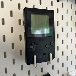May include: A black Game Boy handheld console mounted on a white pegboard. The screen is visible, and the buttons and directional pad are prominent. The console is held in place by a white bracket. Green scissors are also visible.
