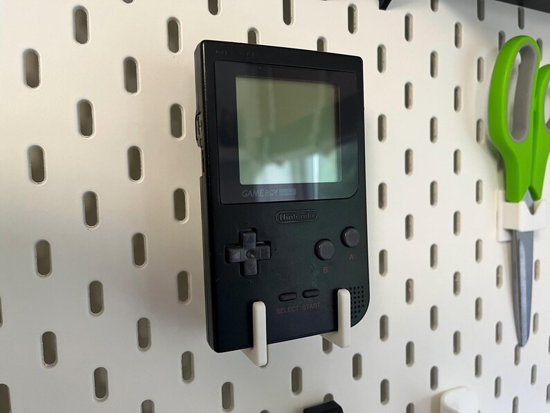 May include: A black Game Boy handheld console mounted on a white pegboard. The screen is visible, and the buttons and directional pad are prominent. The console is held in place by a white bracket. Green scissors are also visible.