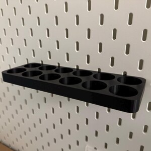 May include: A black organizer with multiple circular holes, designed to attach to a white pegboard. The organizer is rectangular and appears to be made of plastic. The pegboard has a grid of evenly spaced holes.