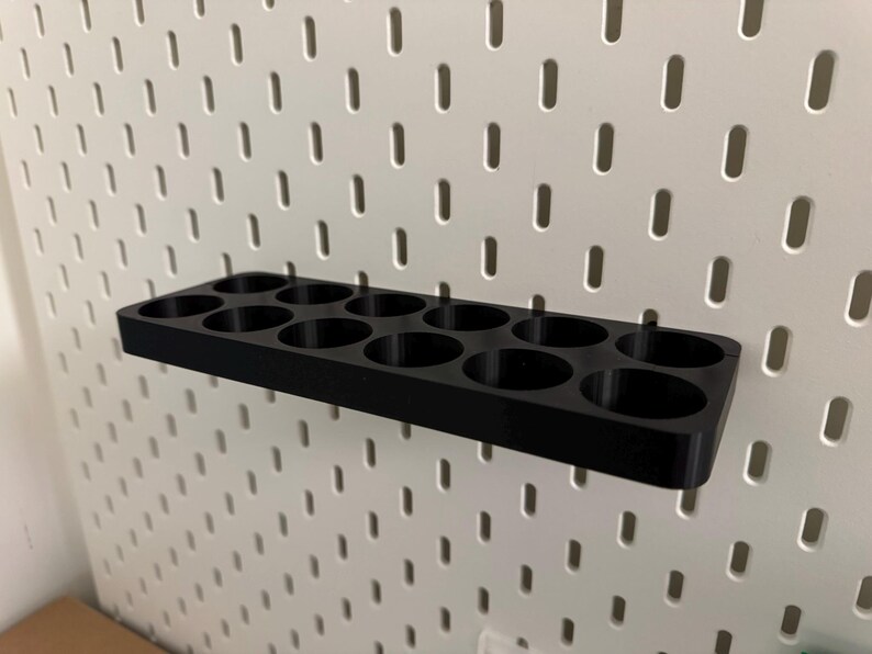 May include: A black organizer with multiple circular holes, designed to attach to a white pegboard. The organizer is rectangular and appears to be made of plastic. The pegboard has a grid of evenly spaced holes.
