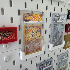 Może przedstawiać: Biała tablica z otworami, na kt&oacute;rej wyświetlane są r&oacute;żne karty i kasety z grami Pokemon. Karty znajdują się w ochronnych plastikowych etui i są zamontowane na tablicy za pomocą przezroczystych plastikowych uchwyt&oacute;w. Karty przedstawiają obrazy postaci Pokemon, w tym Charizarda, i zawierają tekst, kt&oacute;ry zawiera nazwę Pokemon, typ i siłę ataku. Kasety z grami są czerwone i czarne i mają logo Pokemon.