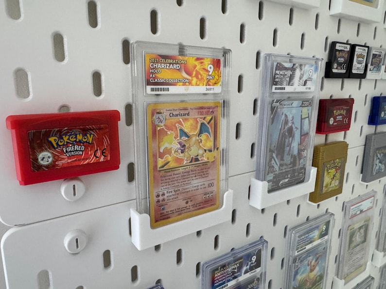 Może przedstawiać: Biała tablica z otworami, na kt&oacute;rej wyświetlane są r&oacute;żne karty i kasety z grami Pokemon. Karty znajdują się w ochronnych plastikowych etui i są zamontowane na tablicy za pomocą przezroczystych plastikowych uchwyt&oacute;w. Karty przedstawiają obrazy postaci Pokemon, w tym Charizarda, i zawierają tekst, kt&oacute;ry zawiera nazwę Pokemon, typ i siłę ataku. Kasety z grami są czerwone i czarne i mają logo Pokemon.