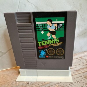 K&ouml;nnte beinhalten: Eine graue Nintendo Entertainment System-Spielkassette f&uuml;r das Spiel "Tennis". Das Etikett zeigt einen pixeligen Tennisspieler in Blau und Schwarz, mit dem Wort "Tennis" in Gr&uuml;n und dem Nintendo-Logo.