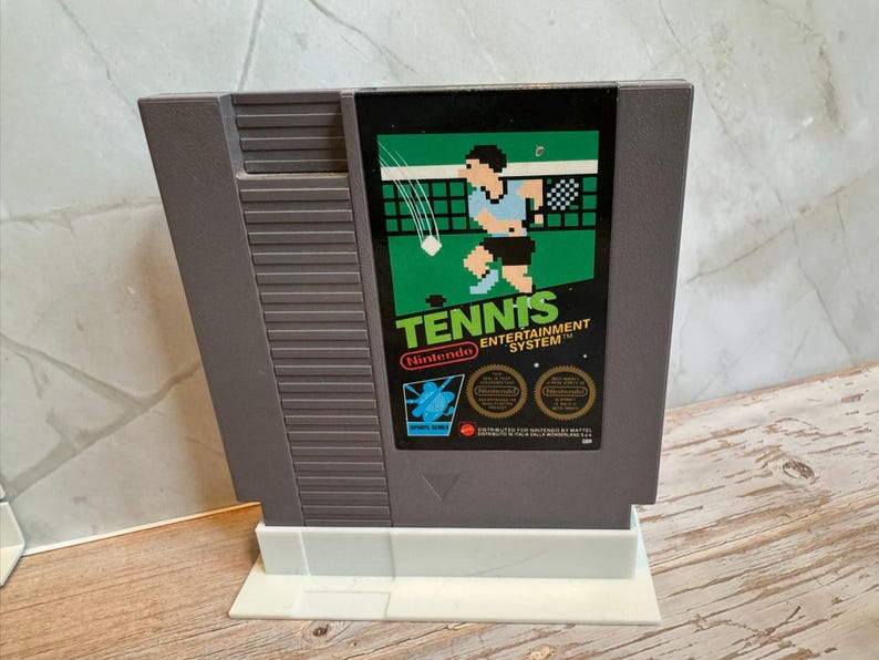 K&ouml;nnte beinhalten: Eine graue Nintendo Entertainment System-Spielkassette f&uuml;r das Spiel "Tennis". Das Etikett zeigt einen pixeligen Tennisspieler in Blau und Schwarz, mit dem Wort "Tennis" in Gr&uuml;n und dem Nintendo-Logo.