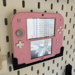 May include: A pink handheld gaming console with a screen displaying language selection options. The console is mounted on a black bracket against a white pegboard wall. The screen shows the text "Select a language."