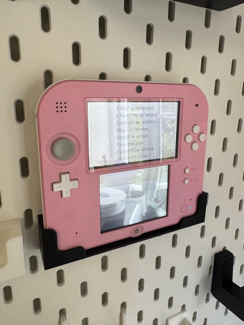 May include: A pink handheld gaming console with a screen displaying language selection options. The console is mounted on a black bracket against a white pegboard wall. The screen shows the text "Select a language."