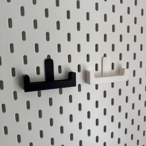 May include: A white pegboard with evenly spaced oval holes. Two shelf brackets are attached. One bracket is black, the other is white. The brackets are designed to hold shelves or other accessories. The pegboard provides a versatile storage solution.