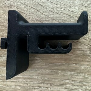 Pu&ograve; includere: Un supporto per cuffie nero stampato in 3D. Il supporto ha una base a forma di T con una parte superiore curva per tenere le cuffie e tre piccole fessure curve. Il supporto &egrave; realizzato in plastica nera e ha una superficie strutturata.