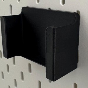 May include: Black plastic wall mount holder with a curved design. The holder is mounted on a white pegboard.
