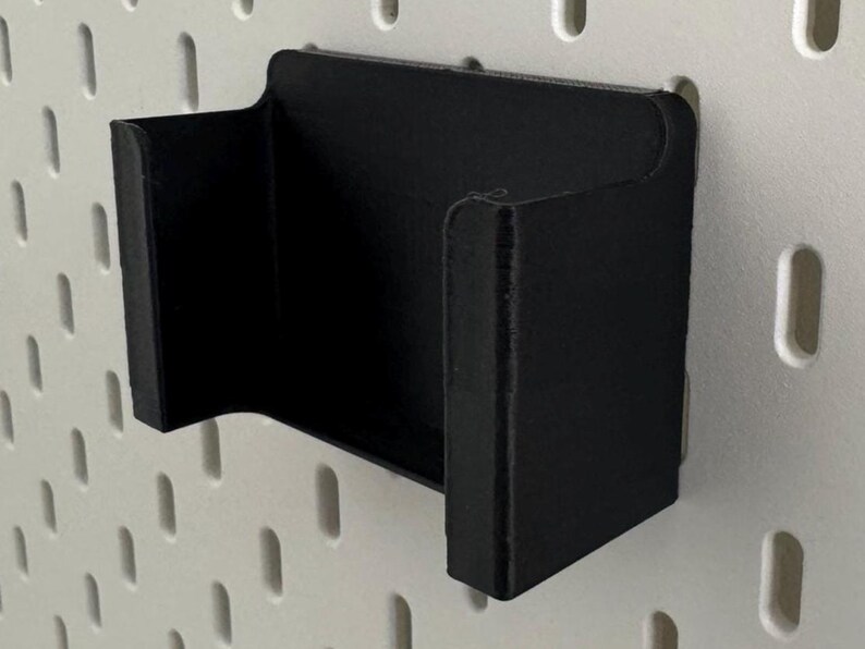 May include: Black plastic wall mount holder with a curved design. The holder is mounted on a white pegboard.
