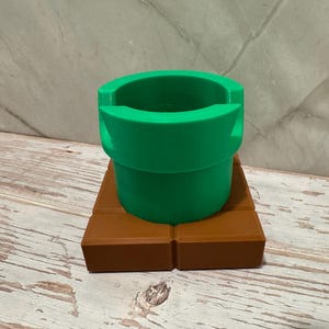 May include: A green pipe, resembling a video game element, is placed on a brown rectangular base. The pipe is cylindrical with a broad opening. The base is made of two rectangular blocks. The design has a playful, cartoonish aesthetic, reminiscent of a video game.