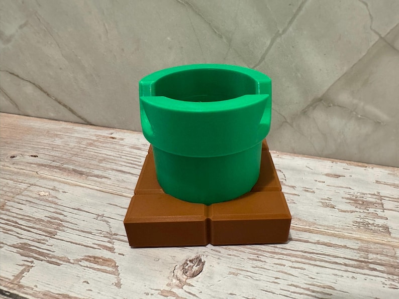 May include: A green pipe, resembling a video game element, is placed on a brown rectangular base. The pipe is cylindrical with a broad opening. The base is made of two rectangular blocks. The design has a playful, cartoonish aesthetic, reminiscent of a video game.