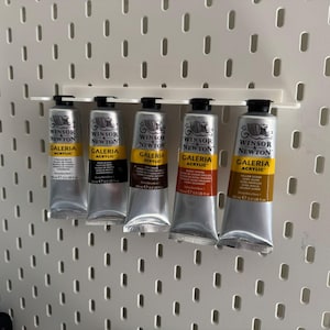May include: Five tubes of Winsor & Newton Galeria acrylic paint in silver tubes, displayed on a white pegboard. The colours include white, black, brown, red-orange, and yellow-orange. Each tube is labelled with the brand and paint type.