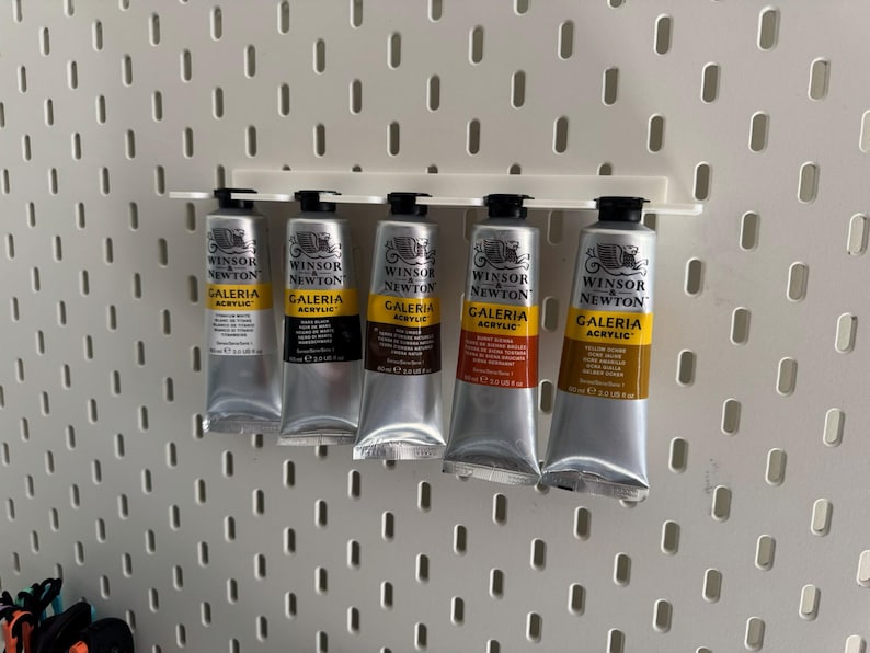 May include: Five tubes of Winsor & Newton Galeria acrylic paint in silver tubes, displayed on a white pegboard. The colours include white, black, brown, red-orange, and yellow-orange. Each tube is labelled with the brand and paint type.