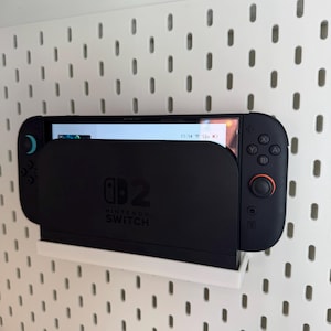 Puede incluir: Una consola Nintendo Switch negra en un soporte blanco, montada en una pared perforada blanca. La consola muestra una pantalla de juego. El soporte tiene el logotipo de Nintendo Switch.