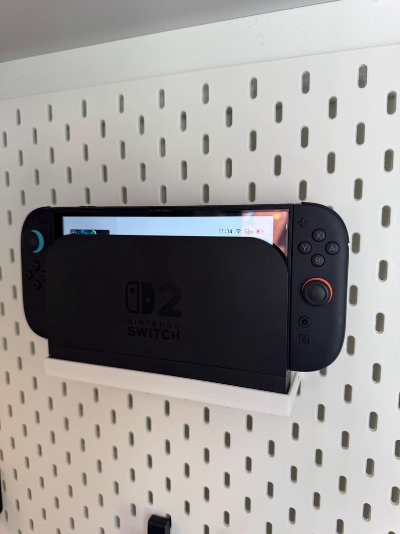 Puede incluir: Una consola Nintendo Switch negra en un soporte blanco, montada en una pared perforada blanca. La consola muestra una pantalla de juego. El soporte tiene el logotipo de Nintendo Switch.