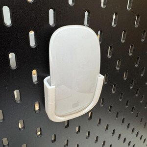 K&ouml;nnte beinhalten: Eine wei&szlig;e Computermaus wird in einem wei&szlig;en, 3D-gedruckten Halter gehalten, der an einer schwarzen Lochwand befestigt ist. Die Maus hat ein glattes, geschwungenes Design und das Apple-Logo ist sichtbar. Der Halter ist so konzipiert, dass er die Maus sicher h&auml;lt.