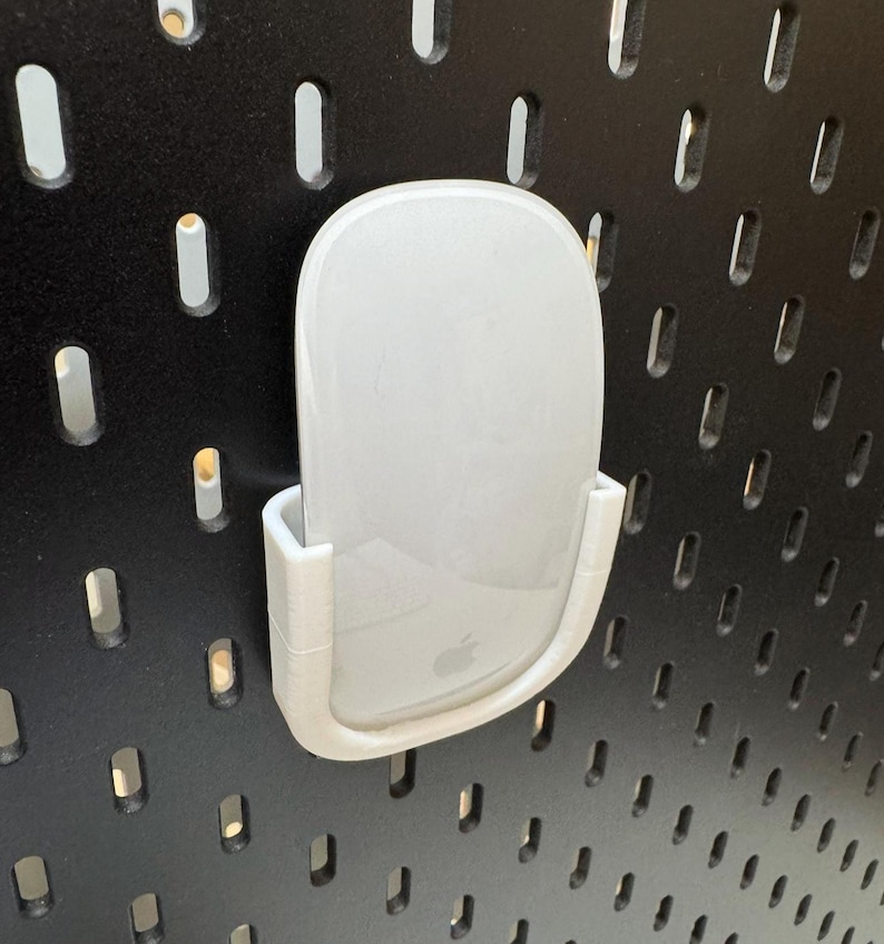 K&ouml;nnte beinhalten: Eine wei&szlig;e Computermaus wird in einem wei&szlig;en, 3D-gedruckten Halter gehalten, der an einer schwarzen Lochwand befestigt ist. Die Maus hat ein glattes, geschwungenes Design und das Apple-Logo ist sichtbar. Der Halter ist so konzipiert, dass er die Maus sicher h&auml;lt.