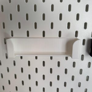 May include: White shelf and black shelf attached to a white pegboard. The white shelf is rectangular with curved edges. The pegboard has evenly spaced oval holes. The black shelf is a small, rectangular holder.