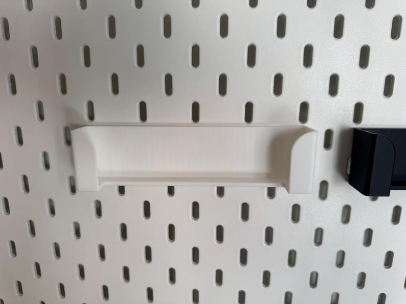 May include: White shelf and black shelf attached to a white pegboard. The white shelf is rectangular with curved edges. The pegboard has evenly spaced oval holes. The black shelf is a small, rectangular holder.