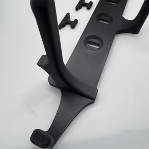 May include: A black 3D-printed device with a curved handle and a series of circular openings. Three small, T-shaped pieces are also present. The device appears to be a holder or mount for an electronic device.