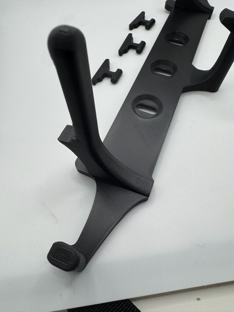 May include: A black 3D-printed device with a curved handle and a series of circular openings. Three small, T-shaped pieces are also present. The device appears to be a holder or mount for an electronic device.