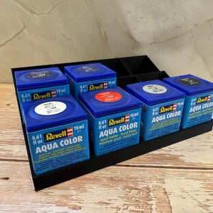 Peut inclure: Un pr&eacute;sentoir noir contient six bo&icirc;tes de peinture Revell Aqua Color bleues. Chaque bo&icirc;te porte le logo Revell, le type de peinture et un volume de 18 ml. Les bo&icirc;tes sont dispos&eacute;es sur une surface en bois.