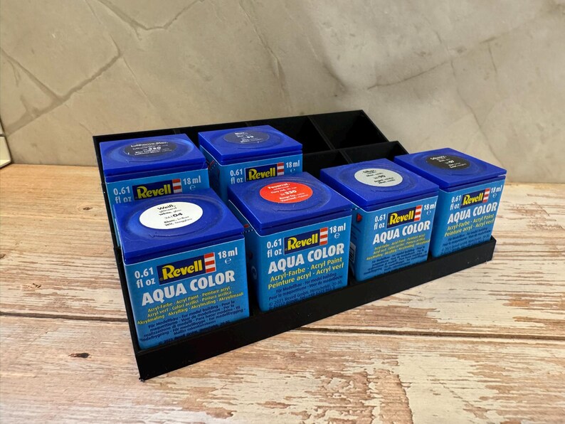 Peut inclure: Un pr&eacute;sentoir noir contient six bo&icirc;tes de peinture Revell Aqua Color bleues. Chaque bo&icirc;te porte le logo Revell, le type de peinture et un volume de 18 ml. Les bo&icirc;tes sont dispos&eacute;es sur une surface en bois.