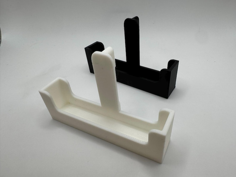 以下が含まれることがあります： 白と黒の2つの3Dプリントされたカードホルダーが白い背景に表示されています。各ホルダーは、中央に垂直のサポートと湾曲したエッジを備えた長方形のベースを持っています。