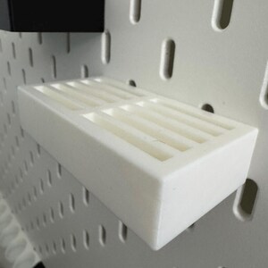 Op de afbeelding: Een wit rechthoekig schap met een sleufvormige bovenkant is gemonteerd op een wit pegboard. Een zwart rechthoekig stuk is boven het schap gemonteerd. Het schap is ontworpen om kleine items te bevatten en is gemaakt van duurzaam materiaal.
