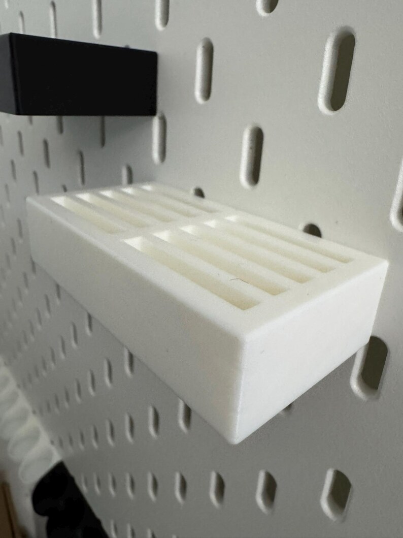 Op de afbeelding: Een wit rechthoekig schap met een sleufvormige bovenkant is gemonteerd op een wit pegboard. Een zwart rechthoekig stuk is boven het schap gemonteerd. Het schap is ontworpen om kleine items te bevatten en is gemaakt van duurzaam materiaal.