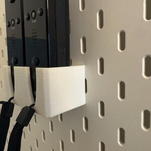 Puede incluir: Un soporte blanco impreso en 3D dise&ntilde;ado para guardar dos mandos Wii negros. El soporte est&aacute; montado en una pared perforada blanca, con las correas de mu&ntilde;eca de los mandos visibles. El dise&ntilde;o es simple y funcional.