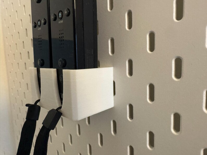 Puede incluir: Un soporte blanco impreso en 3D dise&ntilde;ado para guardar dos mandos Wii negros. El soporte est&aacute; montado en una pared perforada blanca, con las correas de mu&ntilde;eca de los mandos visibles. El dise&ntilde;o es simple y funcional.