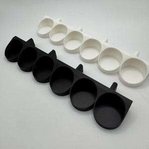 以下が含まれることがあります： 黒と白の2つの3Dプリントオーガナイザー。それぞれに6つの円形コンパートメントがあります。オーガナイザーは、丸みを帯びたエッジと各端の小さなハンドル状の延長部を備えた長方形のベースを備えています。コンパートメントは、小さなアイテムを収納するように設計されています。