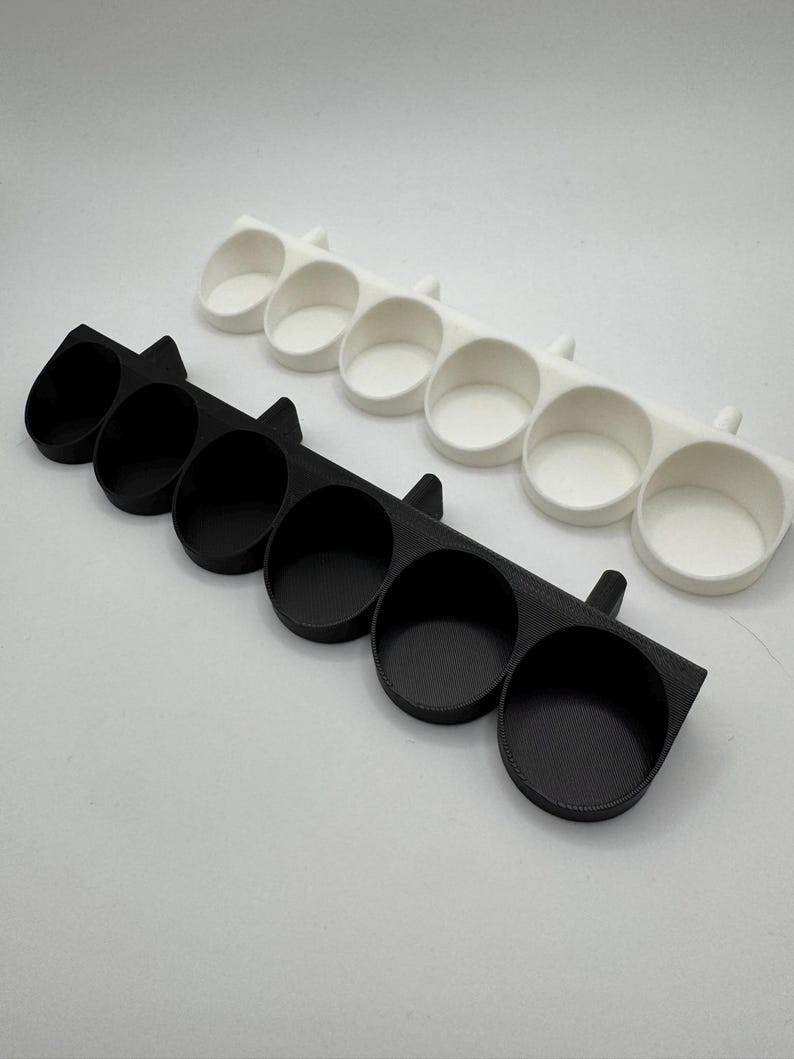 以下が含まれることがあります： 黒と白の2つの3Dプリントオーガナイザー。それぞれに6つの円形コンパートメントがあります。オーガナイザーは、丸みを帯びたエッジと各端の小さなハンドル状の延長部を備えた長方形のベースを備えています。コンパートメントは、小さなアイテムを収納するように設計されています。
