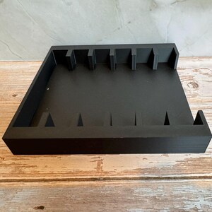 May include: A black wooden organizer with multiple slots and dividers. The organizer has a rectangular shape with raised edges and a series of vertical and triangular cutouts. The item is designed to hold and organize small items.