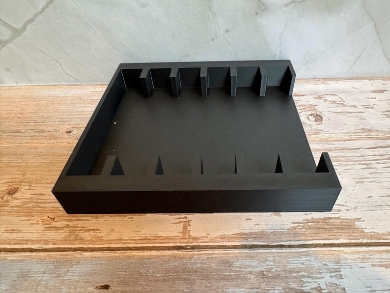 May include: A black wooden organizer with multiple slots and dividers. The organizer has a rectangular shape with raised edges and a series of vertical and triangular cutouts. The item is designed to hold and organize small items.